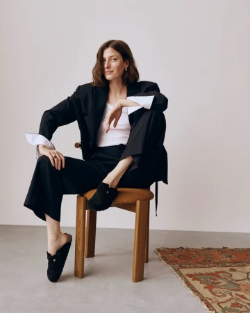 A woman sits on a wooden stool in a black blazer, white top, and wide-legged trousers, posing in an indoor studio setting.