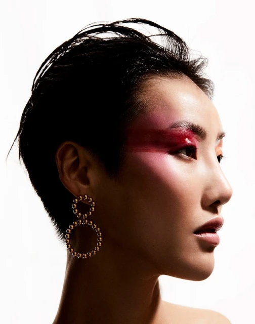 Profile portrait of an Asian woman with short black hair, artistic pink eyeshadow, and circular gold earrings against a white background.