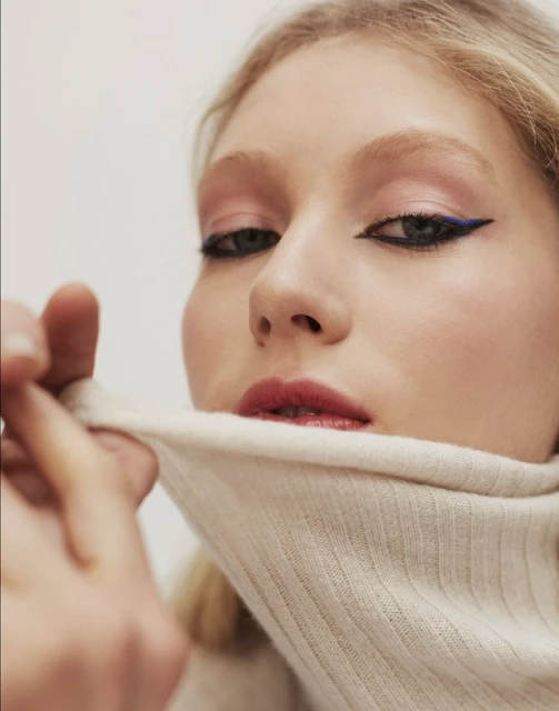 Close-up portrait of an adult woman with fair skin and blonde hair, showcasing makeup with pink eyeshadow and bold blue eyeliner.