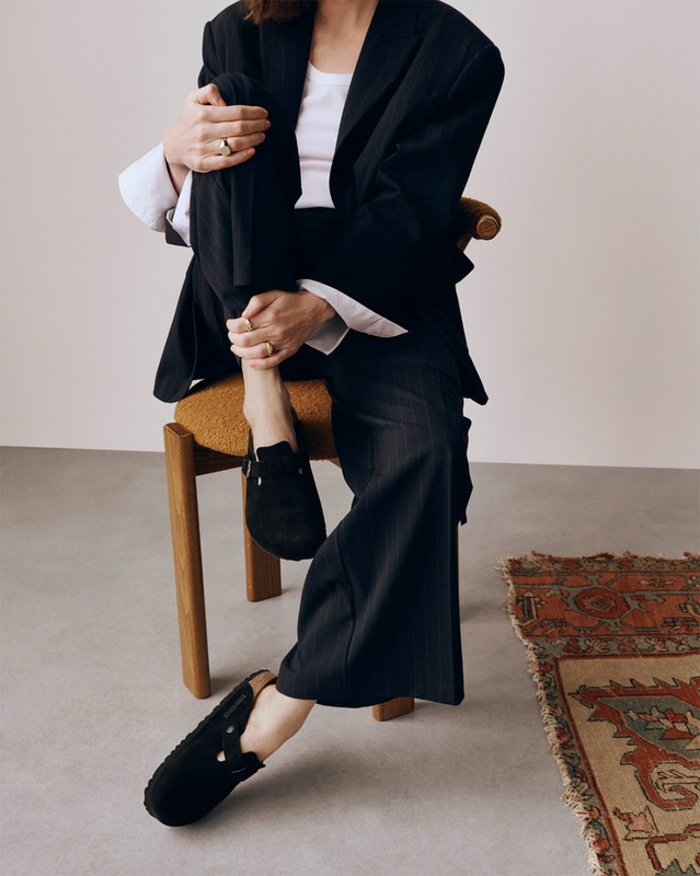 A casually-dressed woman is sitting on a yellow chair with a patterned rug visible in the background in this indoor fashion shot.