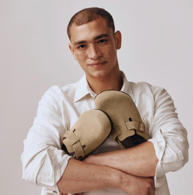 Adult male embraces light brown suede Boston clogs against off-white backdrop in a fashion product shot with a warm, inviting aesthetic.