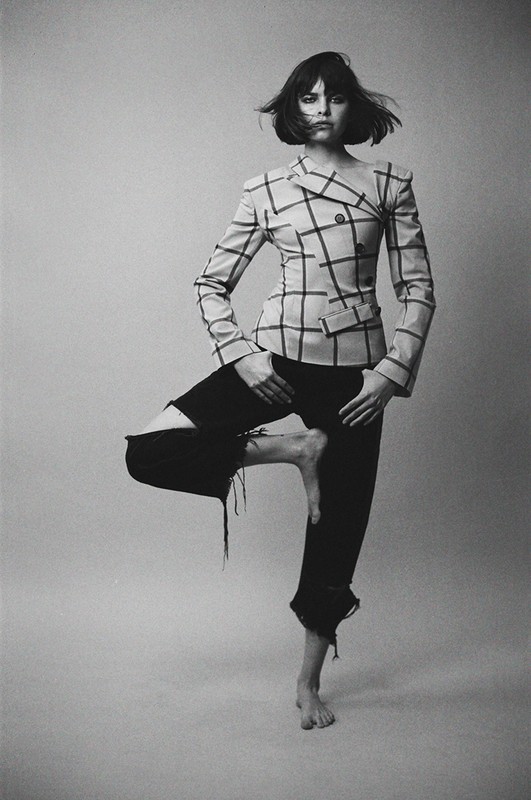 Black and white portrait of a woman posing on one leg wearing a plaid jacket and dark distressed jeans against a light seamless background.