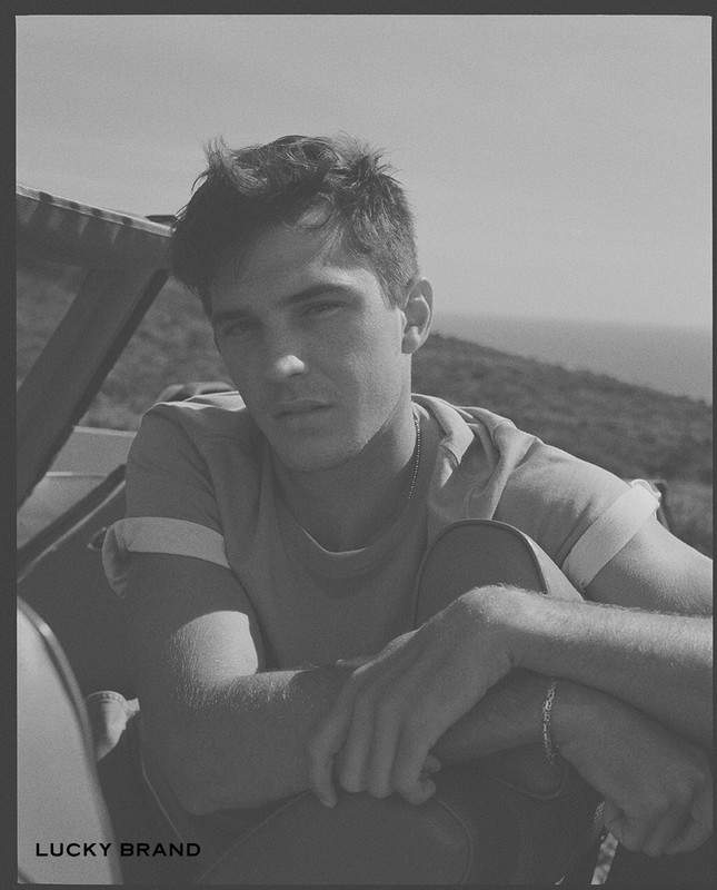 Black and white portrait of a young adult man sitting outside with a light T-shirt and view of a distant mountain range.