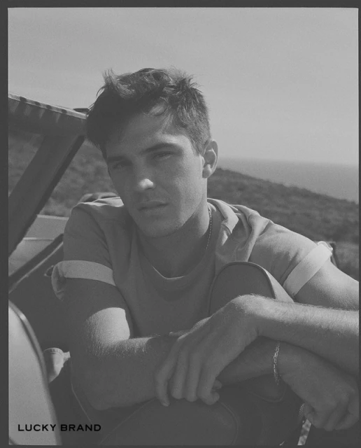 Black and white portrait of a young adult man sitting outside with a light T-shirt and view of a distant mountain range.