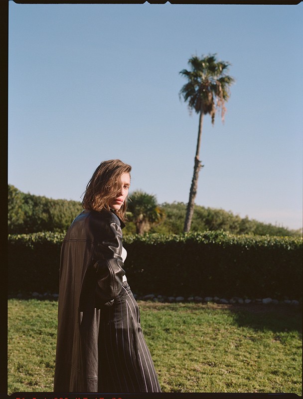 A woman in a dark coat and striped pants stands in a yard with a palm tree in the background under a clear blue sky in daylight.