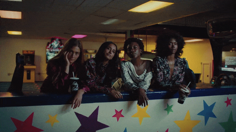 Four young women, diverse ethnicities, seated indoors with drinks, styled in colorful patterns, vintage-inspired fashion, and dark, moody lighting.