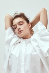Close-up portrait of a woman in a white button-down shirt with her hands clasped behind her head against a plain background
