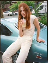 Red-haired woman in cream top and trousers leaning against a vintage blue car drinking from a plastic cup with a straw