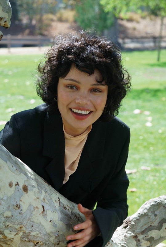 Smiling woman with curly dark hair in a black blazer posing outdoors among tree branches and green foliage, looking directly at the camera.