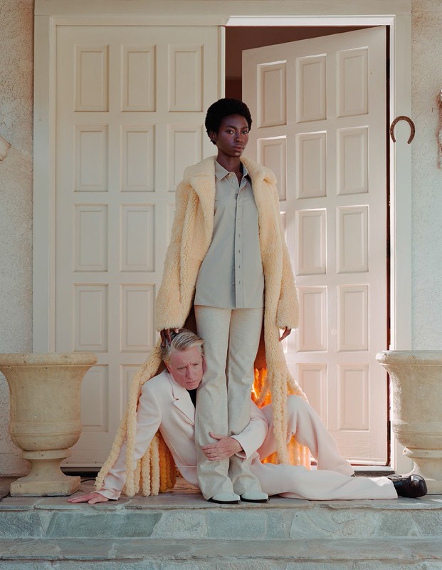 A black female model stands on the legs of a caucasian male model dressed in a tan suit in front of a white door outdoors.
