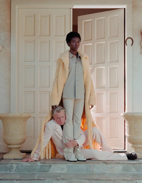 A black female model stands on the legs of a caucasian male model dressed in a tan suit in front of a white door outdoors.