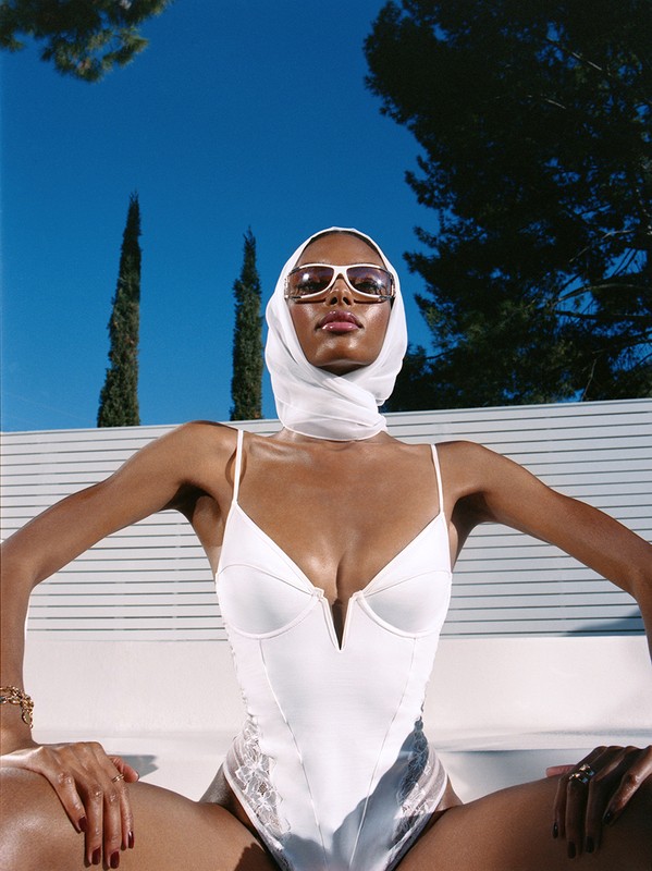 A woman in a white bathing suit and headscarf poses outdoors against a blue sky, displaying fashion and style with natural lighting.