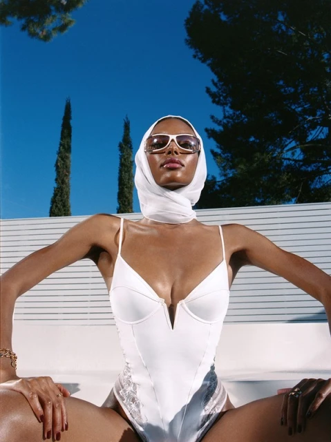 A woman in a white bathing suit and headscarf poses outdoors against a blue sky, displaying fashion and style with natural lighting.