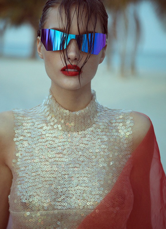 Woman with shiny sunglasses and sequin top posing on a beach; bright red lipstick adds contrast in this fashion-style portrait.