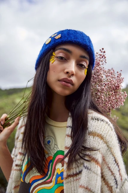 Portrait of a woman with flower petals as makeup and a blue knitted hat, posing outdoors in a patterned sweater with flowers.