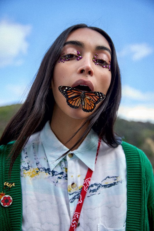 Woman with butterfly on mouth, wearing glitter makeup in outdoor setting with blue sky and hills in the background. 