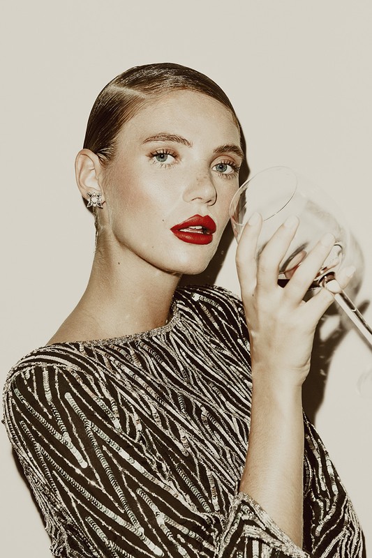 A woman with slicked-back hair and red lipstick holds a wine glass in a beauty style portrait against a neutral background.