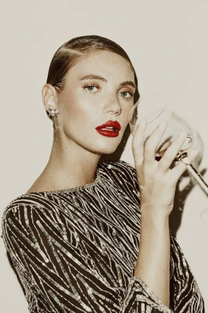 A woman with slicked-back hair and red lipstick holds a wine glass in a beauty style portrait against a neutral background.