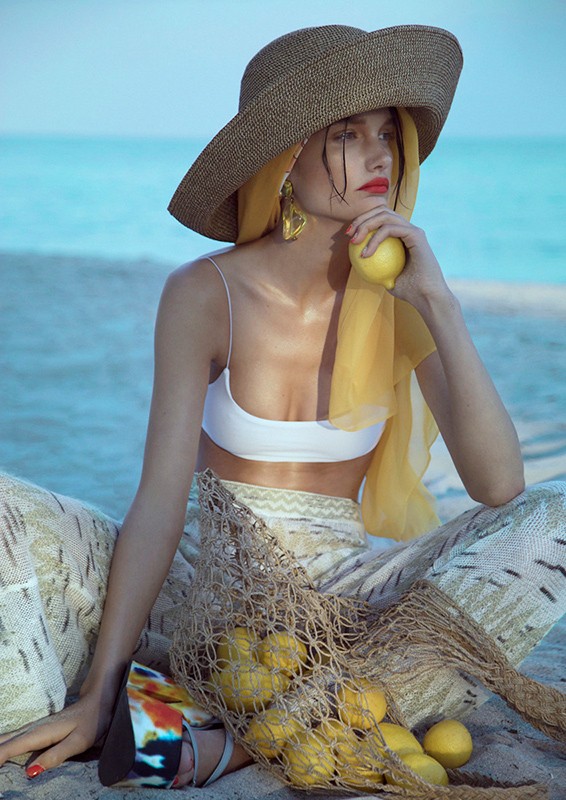 Woman in white bikini top, patterned pants, and straw hat sits on a beach holding a lemon, with a bag of lemons nearby.