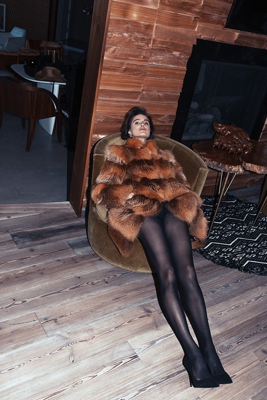 Elegant adult female relaxes in a chair wearing a fur coat, black tights, and heels indoors with a wood paneled wall and floor.