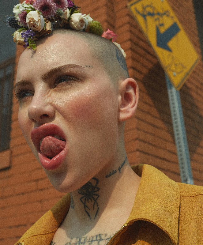 Portrait of a young woman with a shaved head adorned with colorful flowers, sticking out her tongue against a brick wall backdrop.