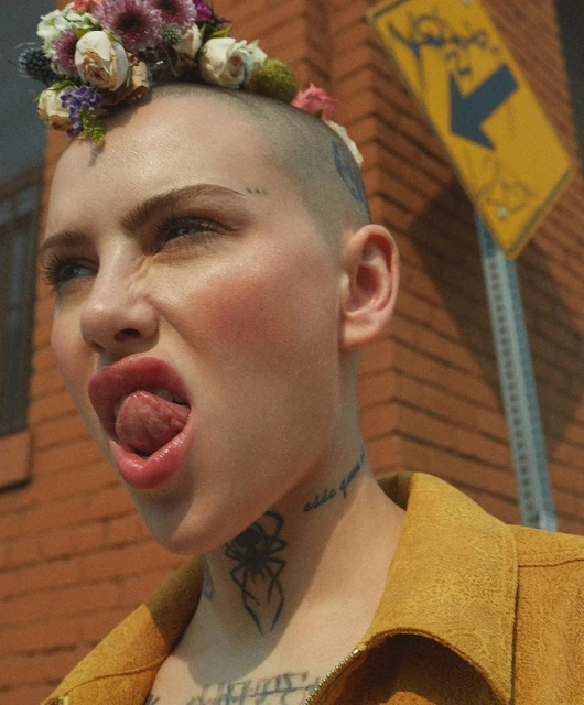 Portrait of a young woman with a shaved head adorned with colorful flowers, sticking out her tongue against a brick wall backdrop.