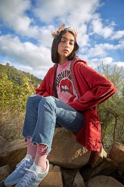 A young woman with a crown and casual clothes sits on a rock outdoors, framed against a blue sky with scattered clouds.