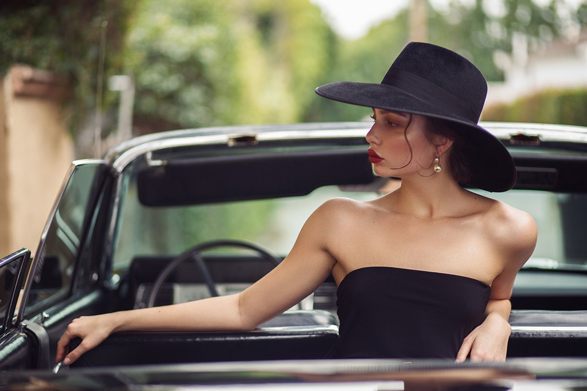 Elegant woman in black hat and dress poses in a classic black convertible car against a blurred background of foliage and buildings.