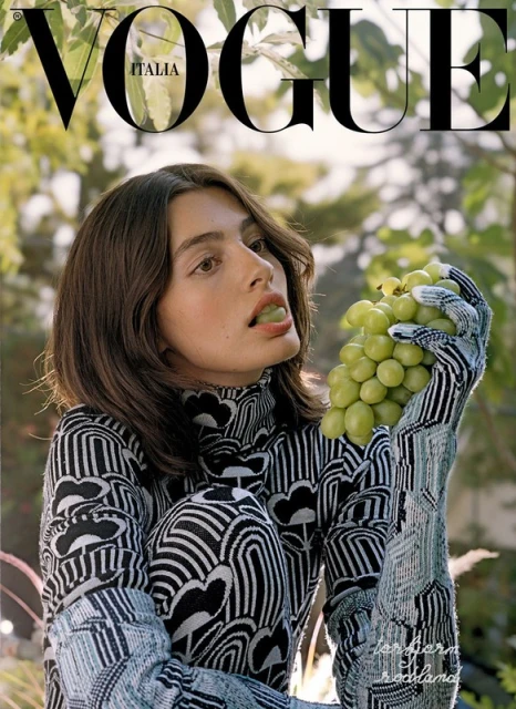 Vogue Italia cover featuring a woman in a patterned turtleneck and gloves eating grapes outdoors, framed by green leaves.