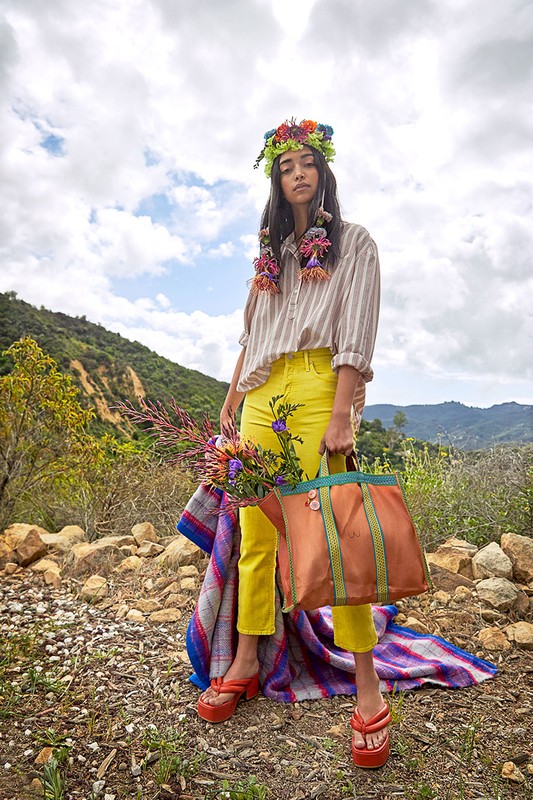 A woman stands on a rocky path in yellow pants and platform sandals, holding a flower bouquet and orange tote bag against a cloudscape.
