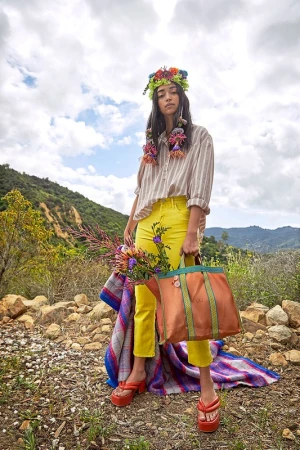 A woman stands on a rocky path in yellow pants and platform sandals, holding a flower bouquet and orange tote bag against a cloudscape.