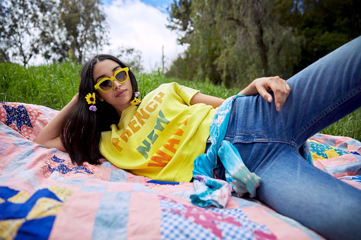 A young woman relaxes on a quilt in a grassy field, wearing a yellow t-shirt and sunglasses. Flowers adorn her hair in a peaceful outdoor scene.