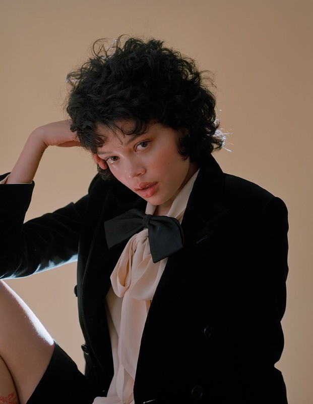 Portrait of a young woman with short curly dark hair, wearing a black blazer and cream colored blouse against a peach background.