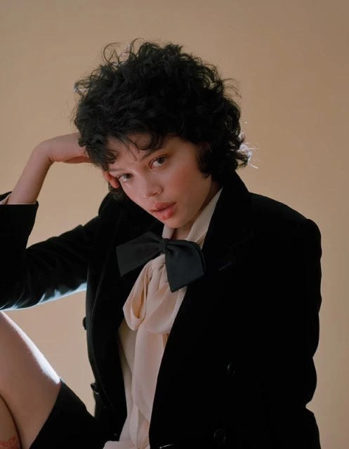 Portrait of a young woman with short curly dark hair, wearing a black blazer and cream colored blouse against a peach background.
