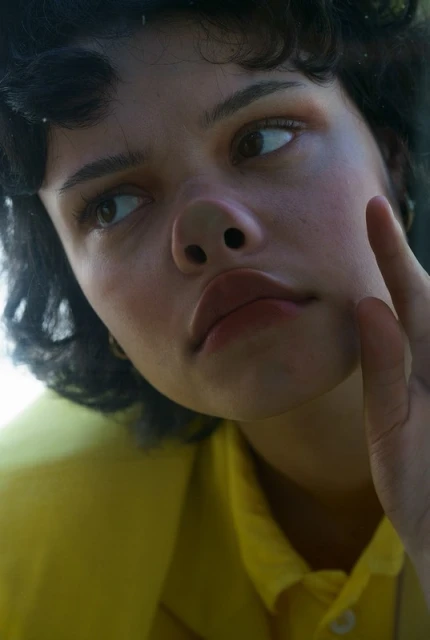Close-up portrait of a brunette woman in a yellow collared shirt, touching her face and looking upwards in a brightly-lit setting.