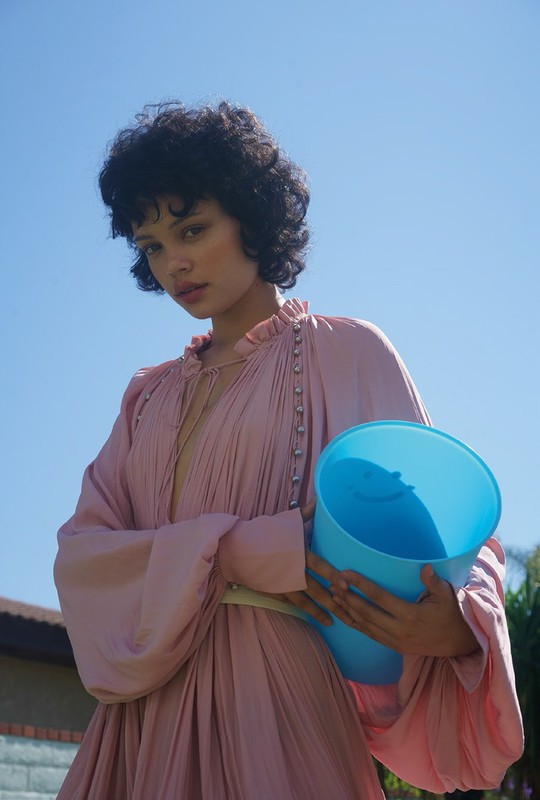 A young woman poses outdoors in a pink dress, holding a light blue plastic bucket, against a clear blue sky with a few trees.