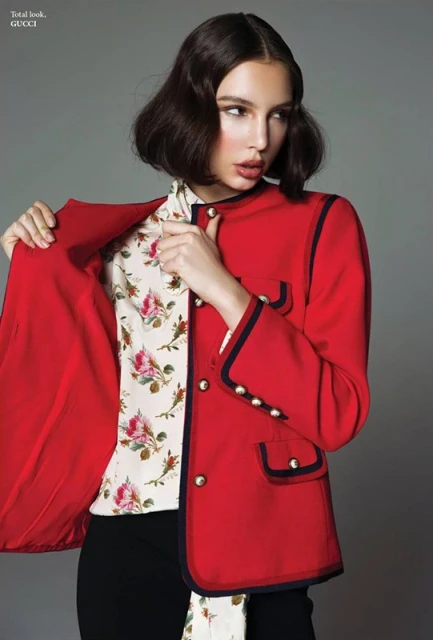Fashion shot of a brunette woman in a red jacket and floral blouse posing against a grey backdrop in a studio environment.
