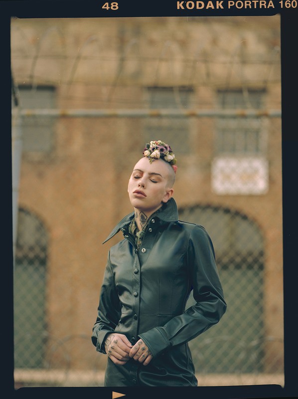 Adult woman in black coat with flower crown, standing outside a building with a fence in the foreground on a cloudy day.