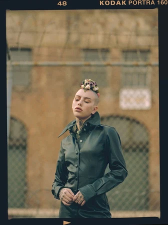 Adult woman in black coat with flower crown, standing outside a building with a fence in the foreground on a cloudy day.