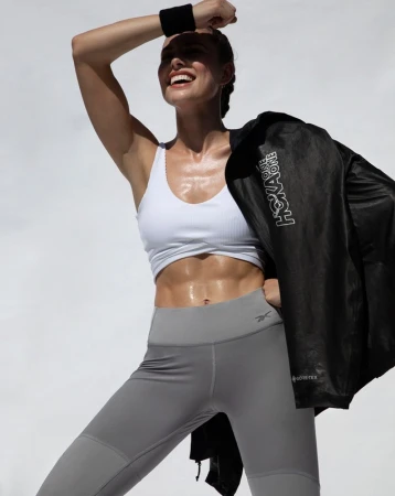Athletic woman smiling and sweating after workout, wearing sportswear and black jacket; fitness concept; indoor setting with neutral background.