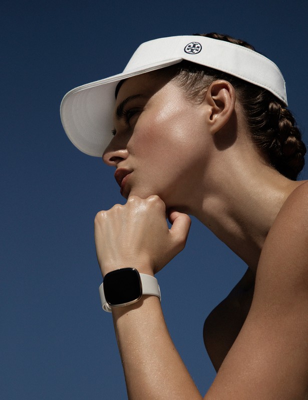 Woman in visor displays smart watch against blue sky. Head and wrist visible, showcasing fashion and technology.