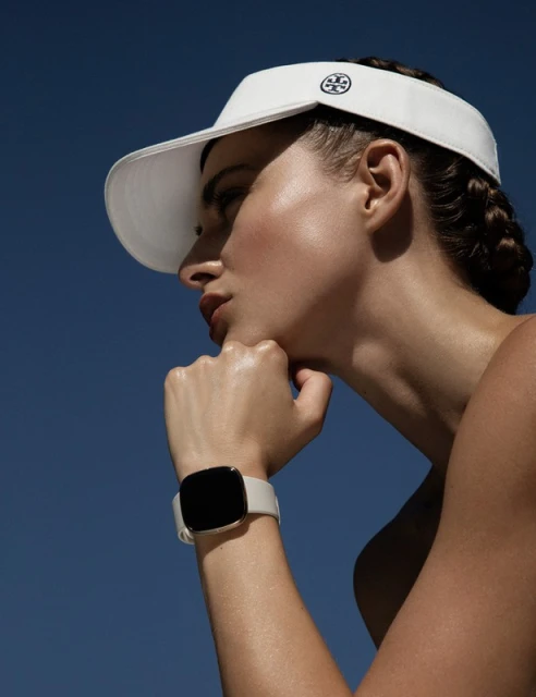 Woman in visor displays smart watch against blue sky. Head and wrist visible, showcasing fashion and technology.