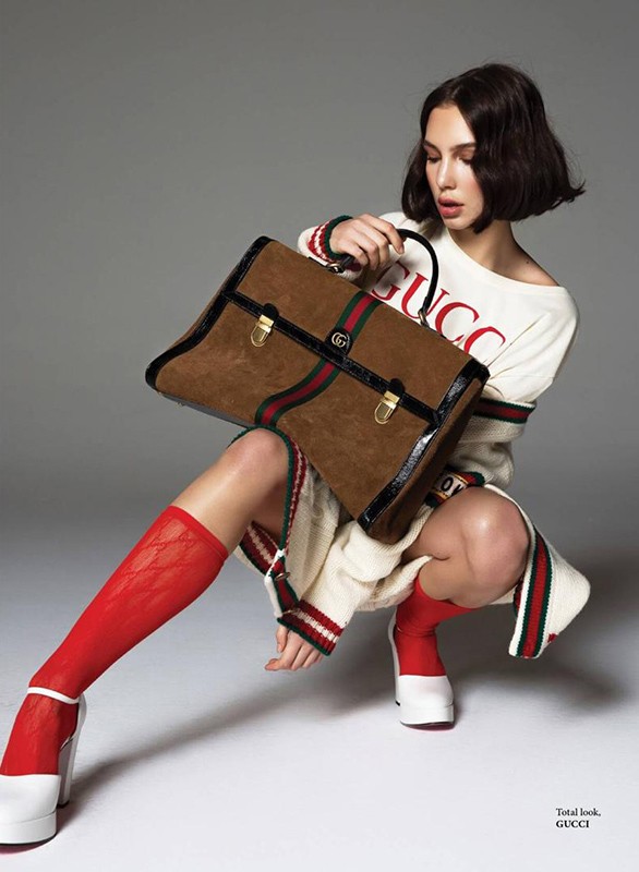 A woman wearing a white sweater and red socks crouches while holding a brown Gucci handbag in a studio shot against a grey background.