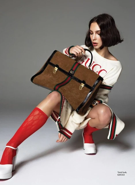 A woman wearing a white sweater and red socks crouches while holding a brown Gucci handbag in a studio shot against a grey background.