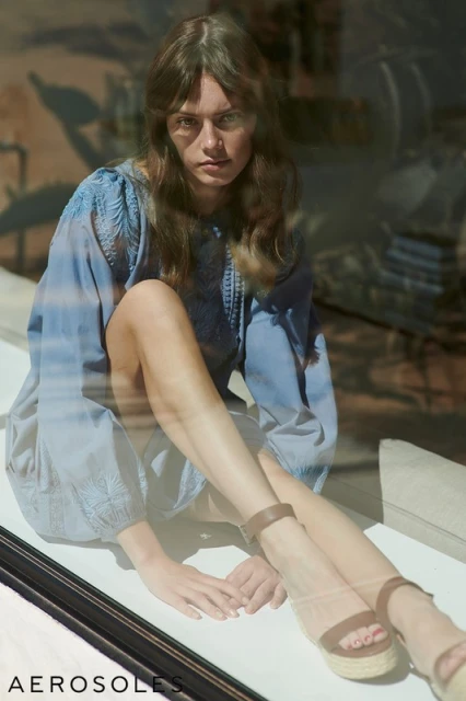 A model sits in a window wearing a light blue tunic dress and brown platform sandals by Aerosoles, glancing at the camera.