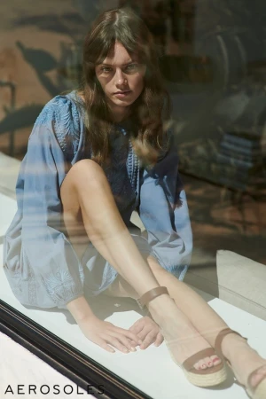 A model sits in a window wearing a light blue tunic dress and brown platform sandals by Aerosoles, glancing at the camera.