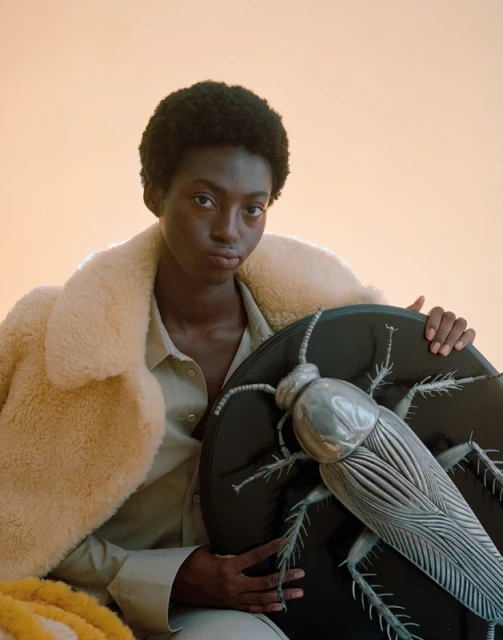 Fashion portrait of a Black woman in a coat holding a large metallic cockroach prop, shot in a brightly lit indoor studio environment.
