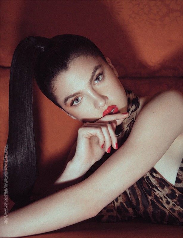 Adult woman with red lipstick poses in front of an orange backdrop, wearing a leopard-print top and her hair in a high ponytail.