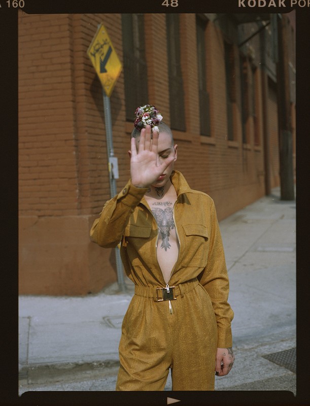 Person with shaved head wearing mustard yellow jumpsuit holding hand up in front of face on Los Angeles street