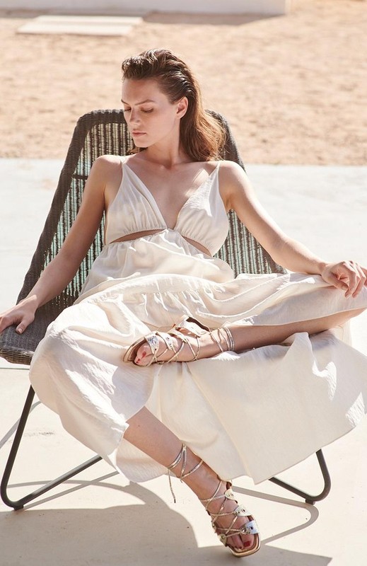 A woman in a white dress and gladiator sandals is seated in a chair outdoors against a neutral background, captured in natural light.